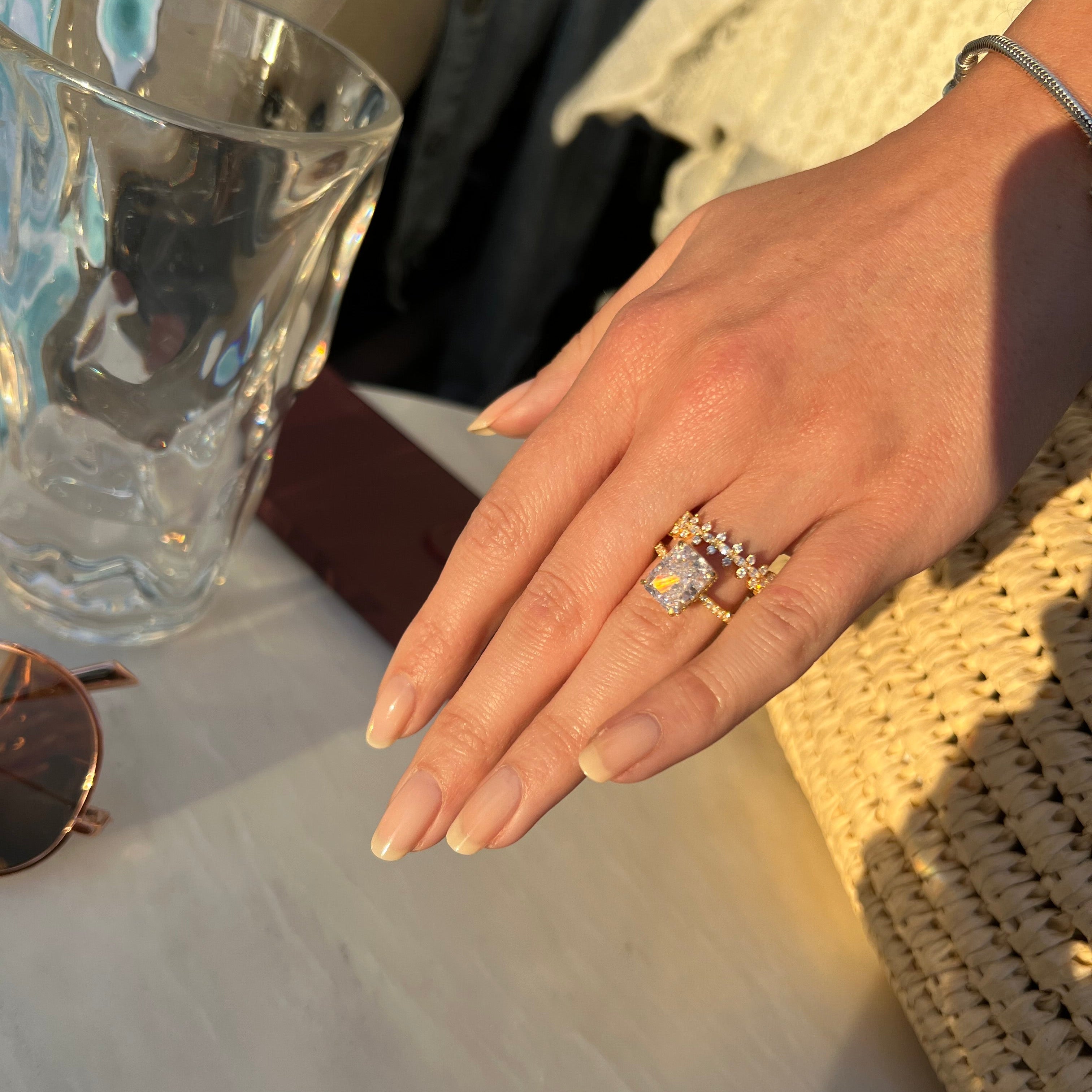 Hand wearing a ring with a clear stone, sitting on a surface with a glass object nearby.