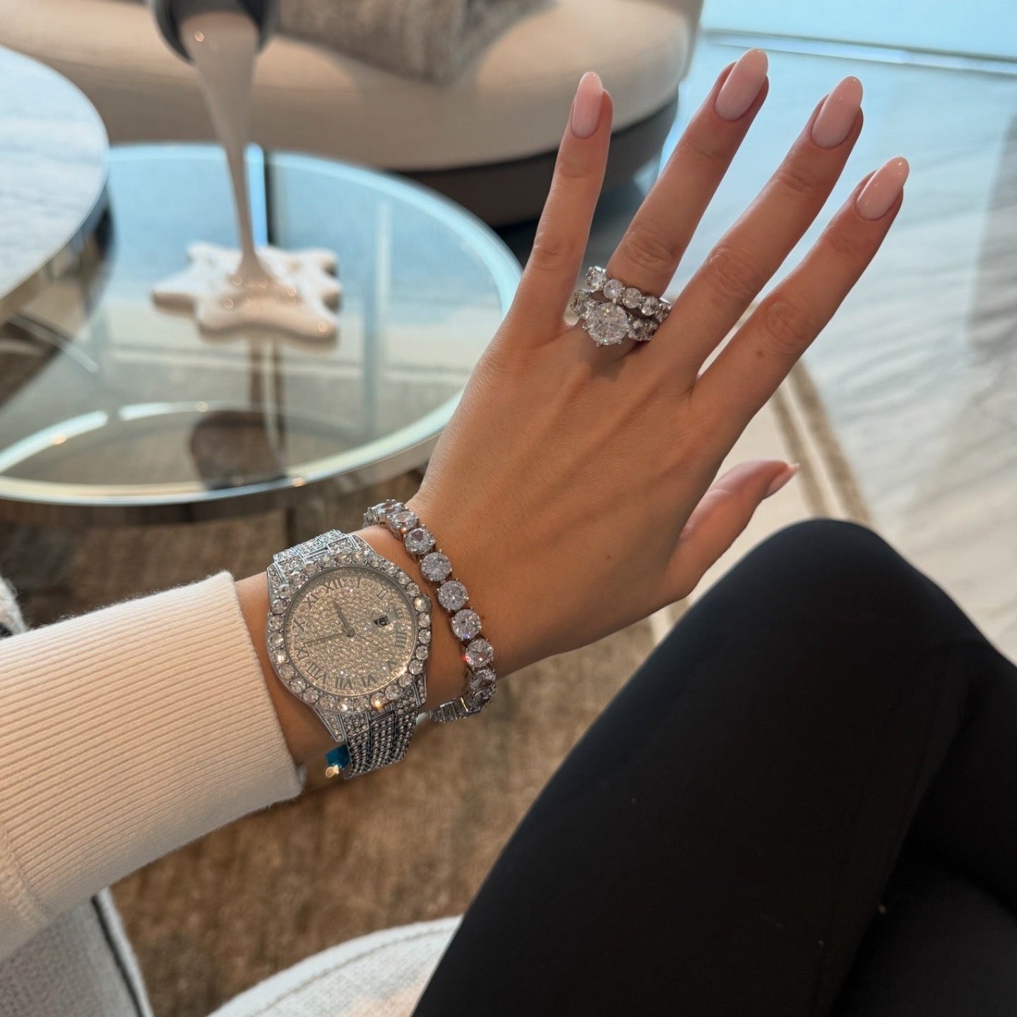 Hand wearing a diamond-studded watch, bracelet, and ring with a blurred indoor background.